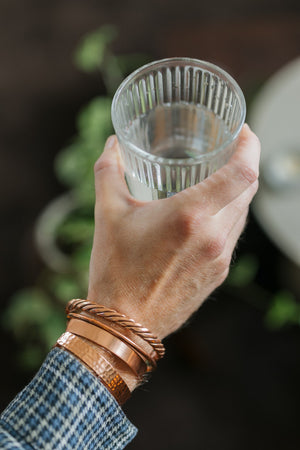 Pulsera de cobre Kopero, las cuatro versiones juntas en una muñeca, con un vaso de agua en la mano