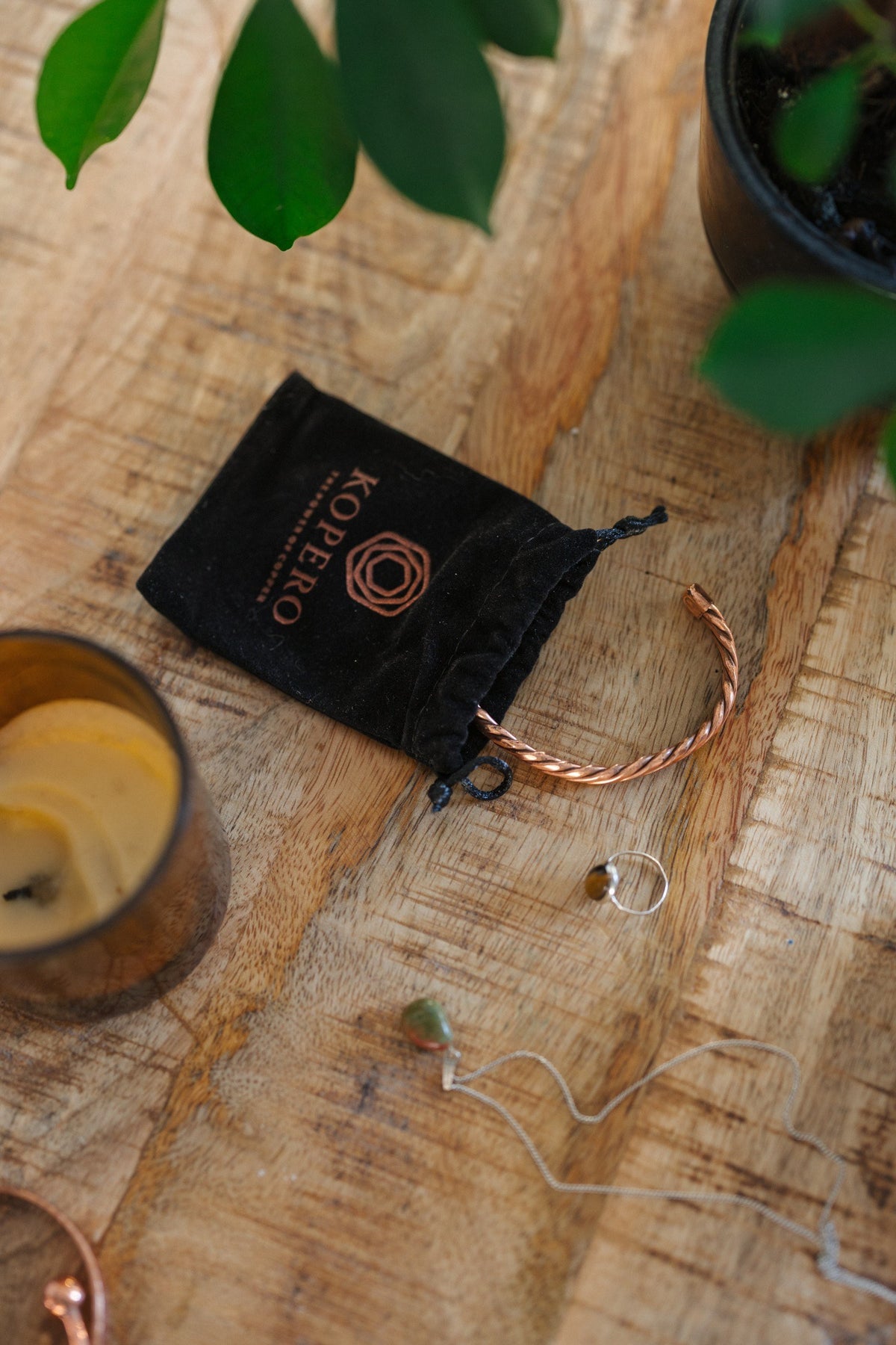 Pulsera de cobre Kopero Trenzado, sobre una mesa de madera junto a la bolsa de terciopelo, una vela y joyas