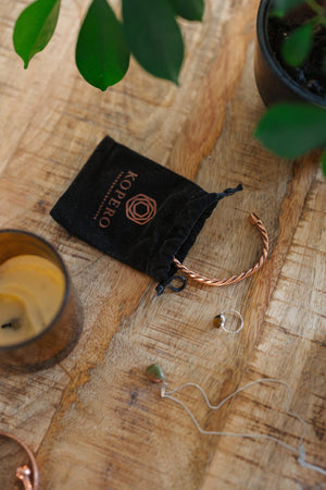 Pulsera de cobre Kopero Trenzado, sobre una mesa de madera junto a la bolsa de terciopelo, una vela y joyas