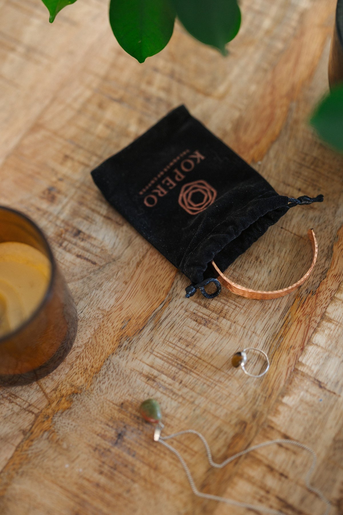 Pulsera de cobre Kopero Martillado, sobre una mesa de madera junto a la bolsa de terciopelo, una vela y joyas