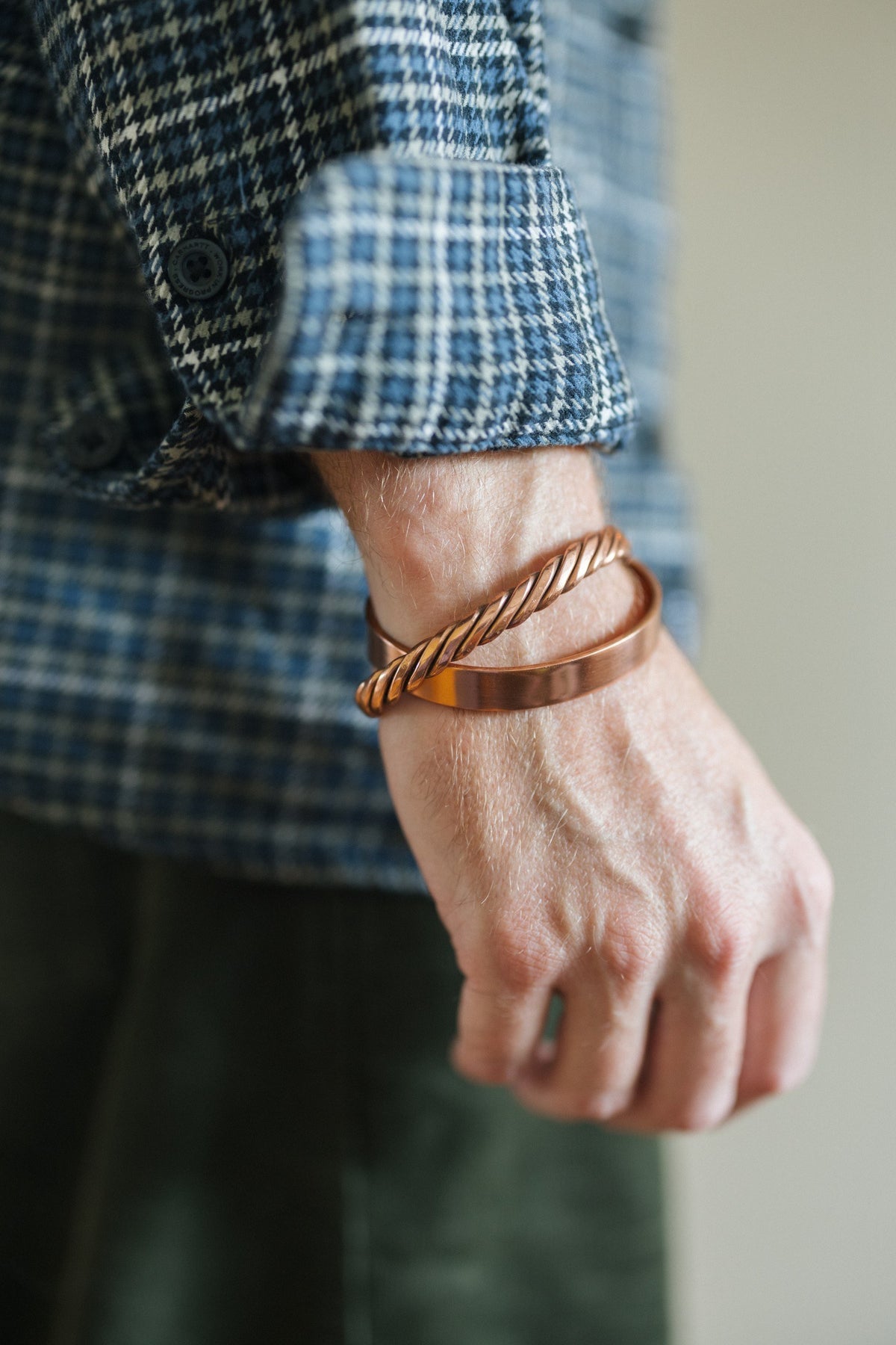 Pulsera de cobre Kopero Liso, en la muñeca sobre una manga de camisa de cuadros, junto con una pulsera trenzada
