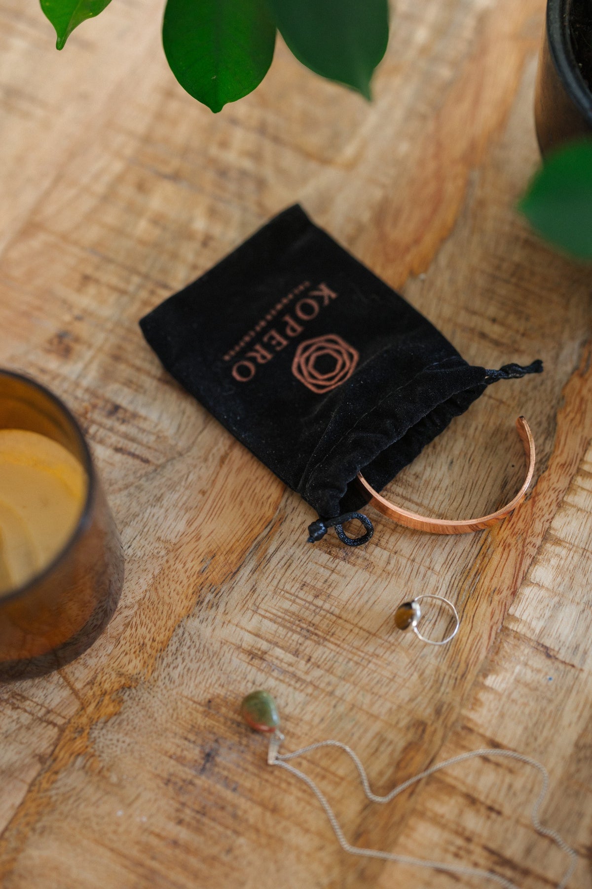 Pulsera de cobre Kopero Liso, sobre una mesa de madera junto a la bolsa de terciopelo, una vela y joyas