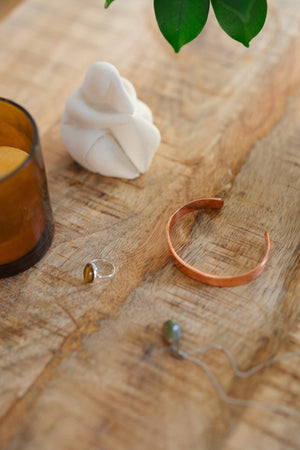 Pulsera de cobre Kopero Liso, sobre una mesa de madera junto a una figura y un anillo