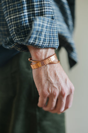 Pulsera de cobre Kopero Redondo, en la muñeca sobre una manga de camisa de cuadros, junto con una pulsera martillada