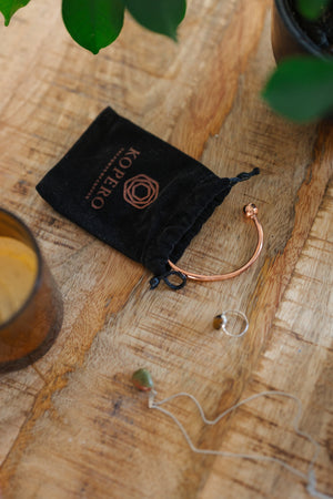 Pulsera de cobre Kopero Redondo, sobre una mesa de madera junto a la bolsa de terciopelo, una vela y joyas