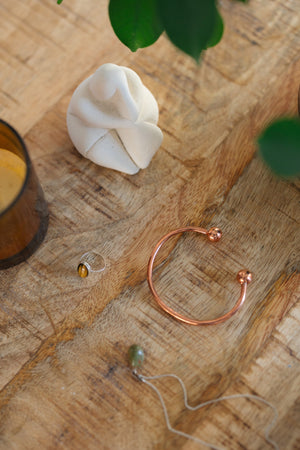Pulsera de cobre Kopero Redondo, sobre una mesa de madera junto a una figura y un anillo