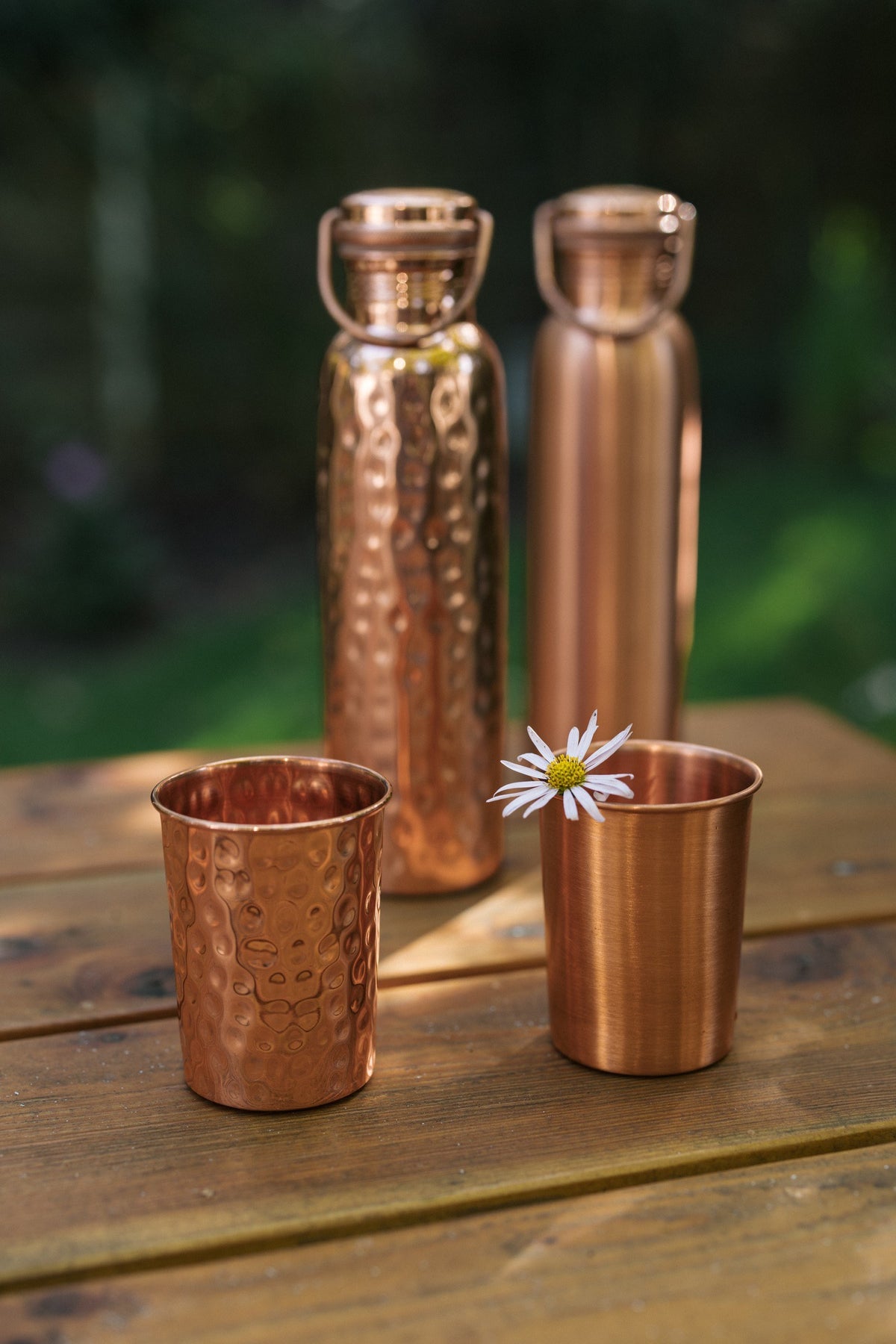 Vaso de cobre Kopero, vasos y botellas lisos y martillados juntos sobre una mesa de madera con una margarita