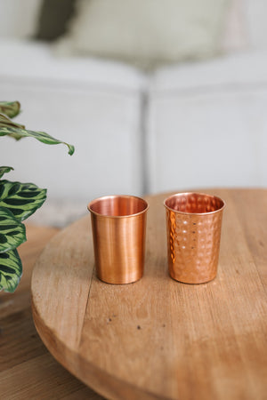 Vaso de cobre Kopero, un vaso liso y uno martillado uno al lado del otro sobre una mesa redonda de madera