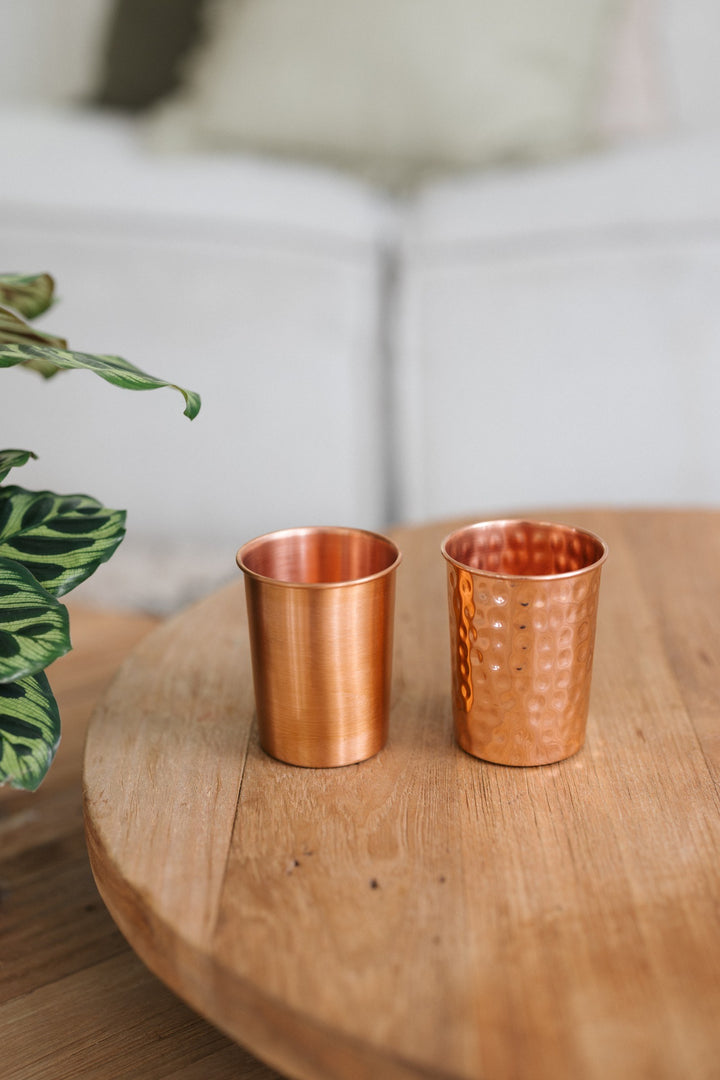 Vaso de cobre Kopero, un vaso liso y uno martillado uno al lado del otro sobre una mesa redonda de madera