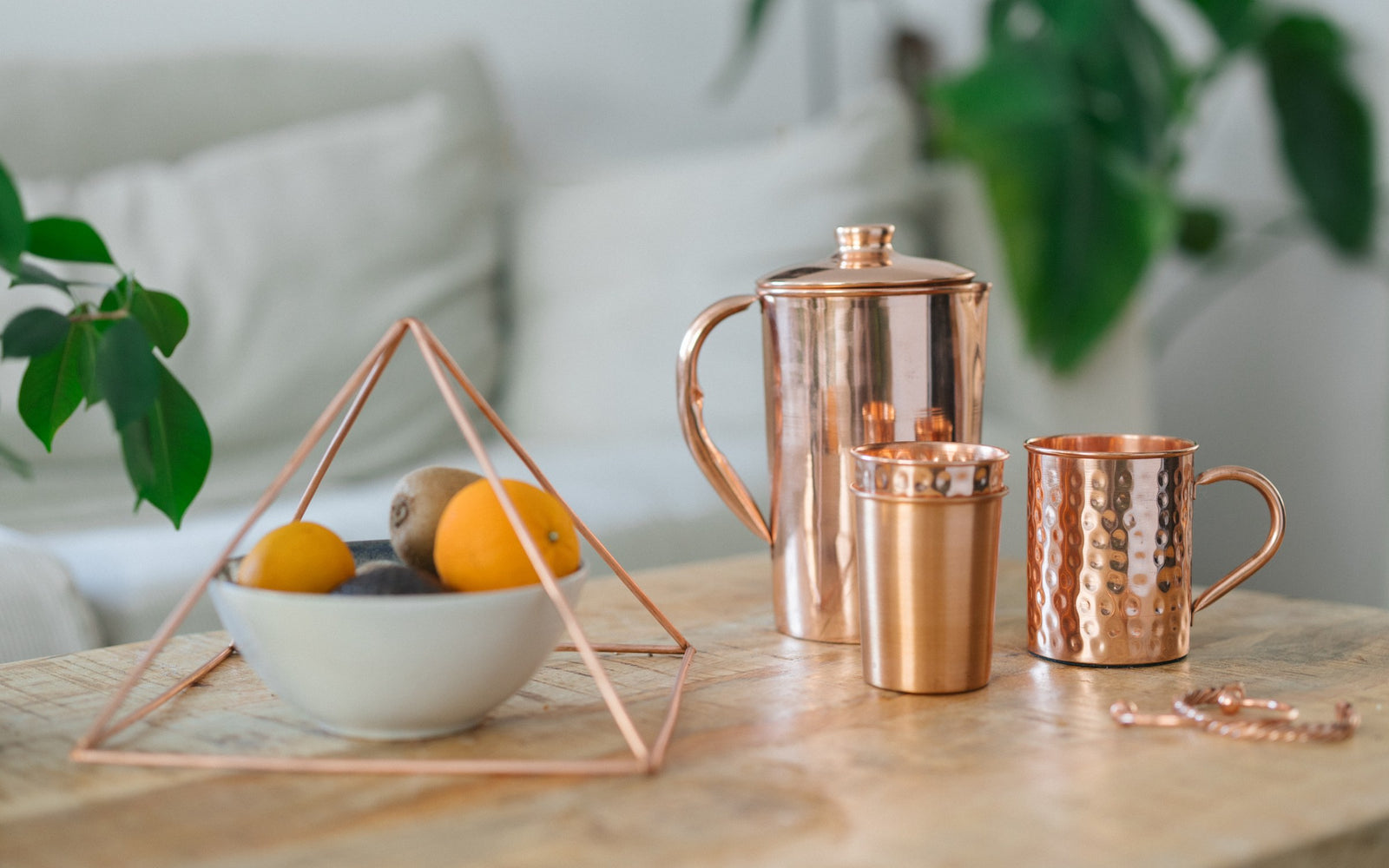 Pirámide de cobre sobre un frutero, con una jarra, un vaso, una taza y una pulsera sobre una mesa de madera