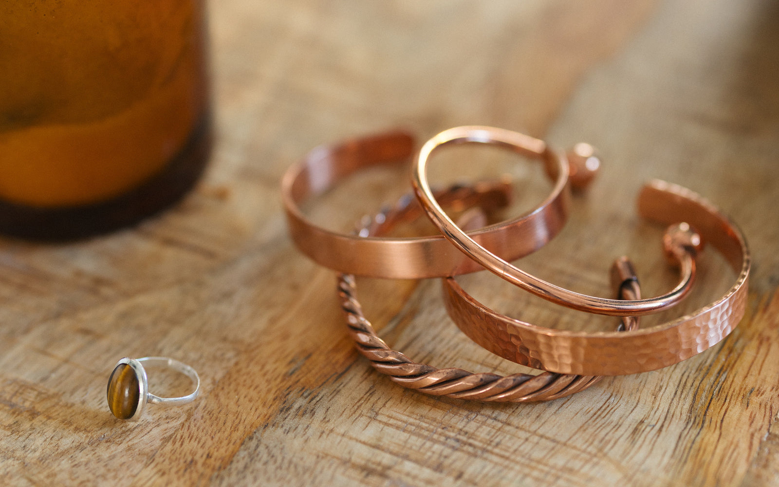 Cuatro pulseras de cobre, trenzada, lisa, martillada y redonda, sobre una mesa de madera junto a una vela y un anillo