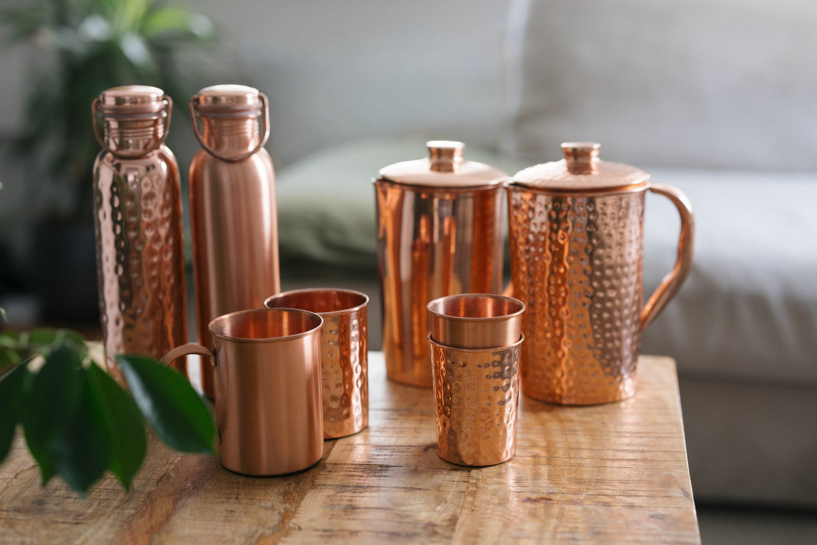 Botellas, jarras, tazas y vasos de cobre sobre una mesa de madera delante de un sofá
