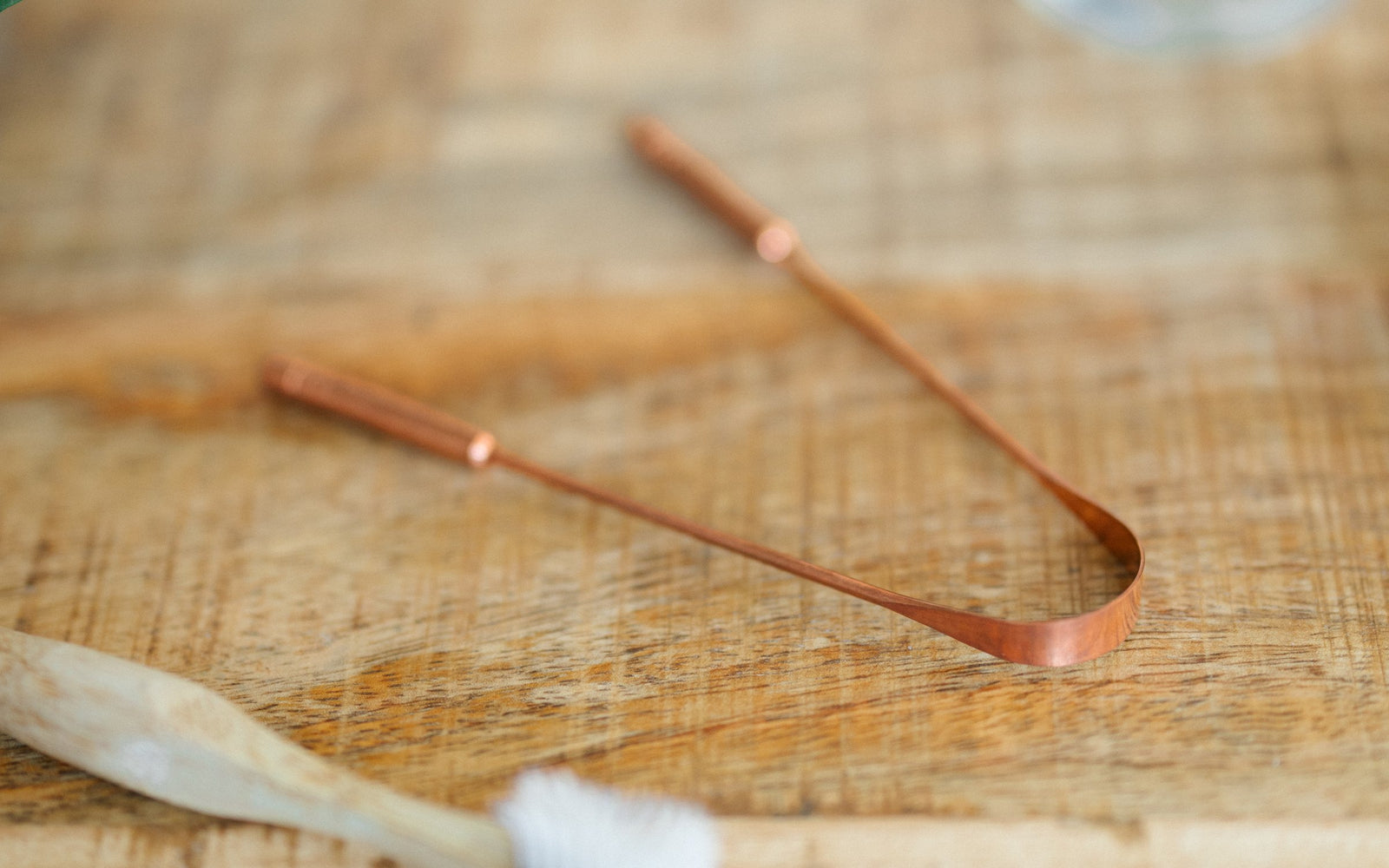 Raspador de lengua de cobre sobre una mesa de madera, junto a un cepillo de dientes de bambú