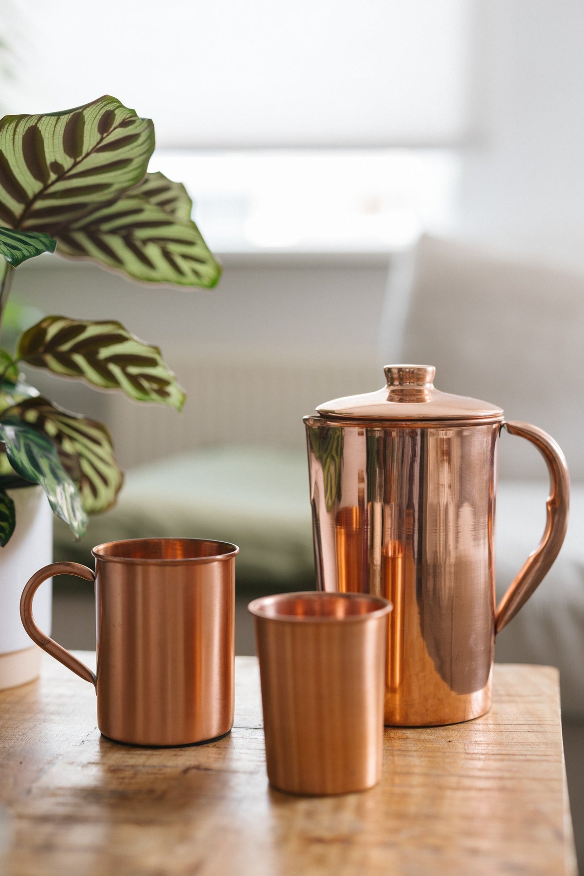 Jarra de cobre Kopero Liso, con una taza y un vaso sobre una mesa de madera