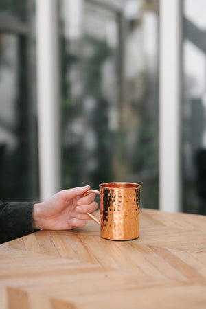 Taza de cobre Kopero Martillado, sobre una mesa de madera, con una mano en el asa