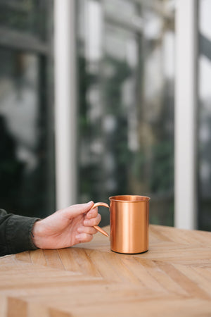 Taza de cobre Kopero Liso, sobre una mesa de madera, con una mano en el asa