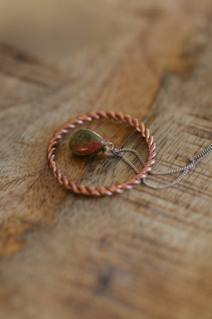 Anillo tensor de cobre Kopero, sobre una mesa de madera con un colgante de unakita