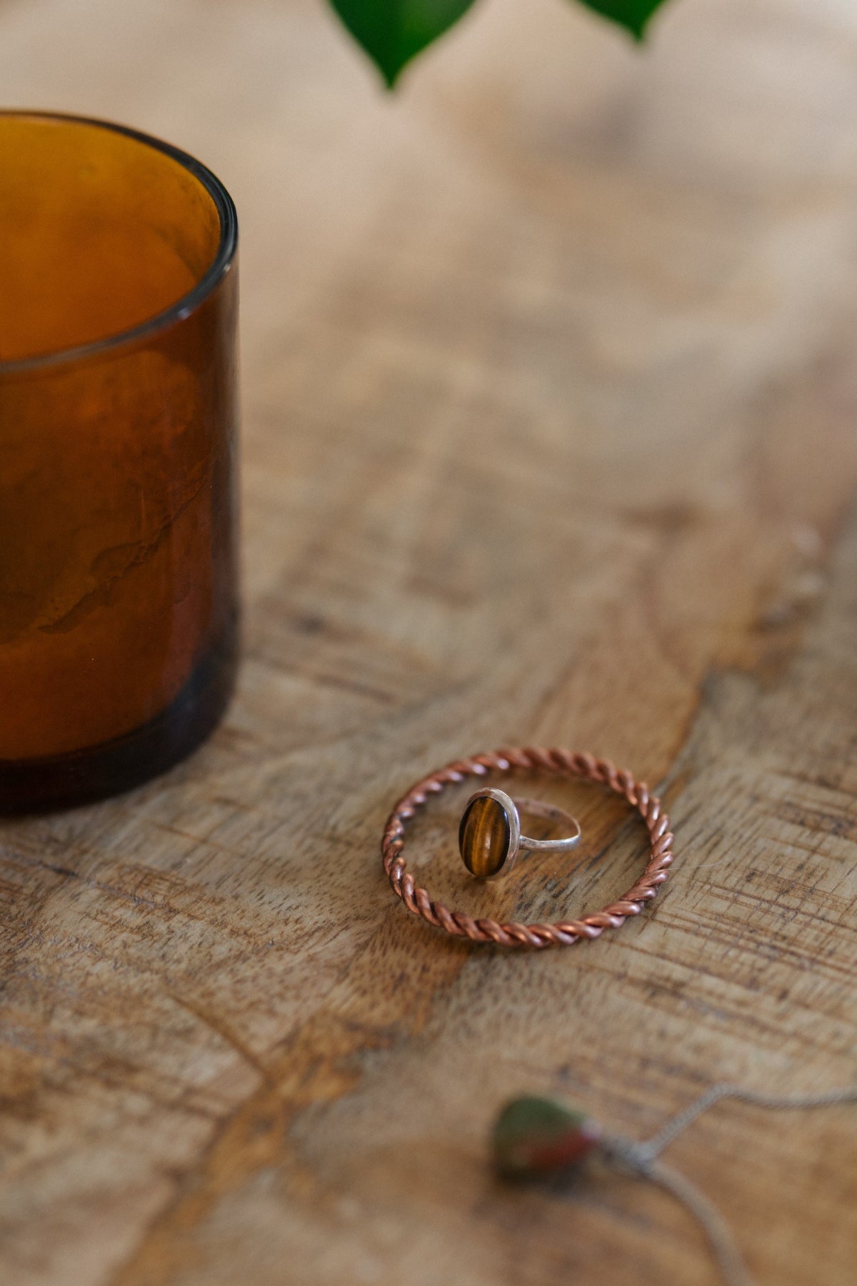 Anillo tensor de cobre Kopero, sobre una mesa de madera con un anillo de ojo de tigre y un portavelas de cristal