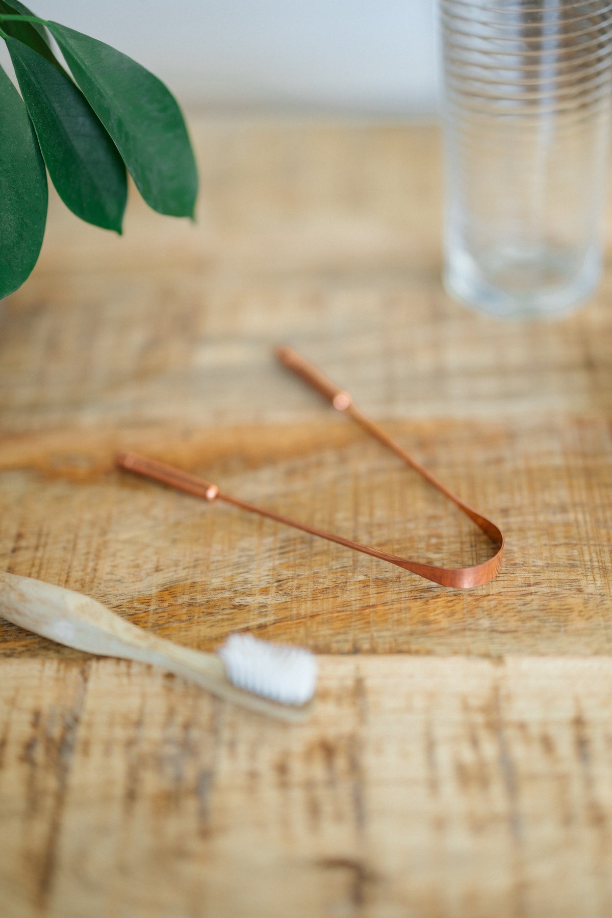 Raspador de lengua de cobre Kopero, sobre una mesa de madera junto a un cepillo de dientes de bambú y un vaso de agua