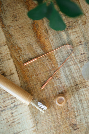 Raspador de lengua de cobre Kopero, sobre una tabla de madera junto a un cepillo de dientes de bambú