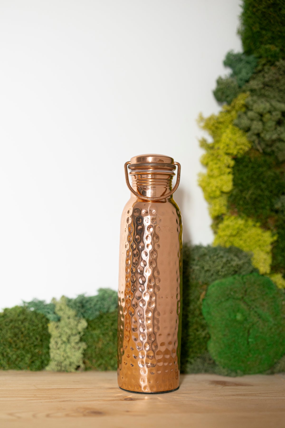 Botella de agua de cobre Kopero Martillada, 900 ml, de pie sobre una mesa de madera con un fondo de musgo