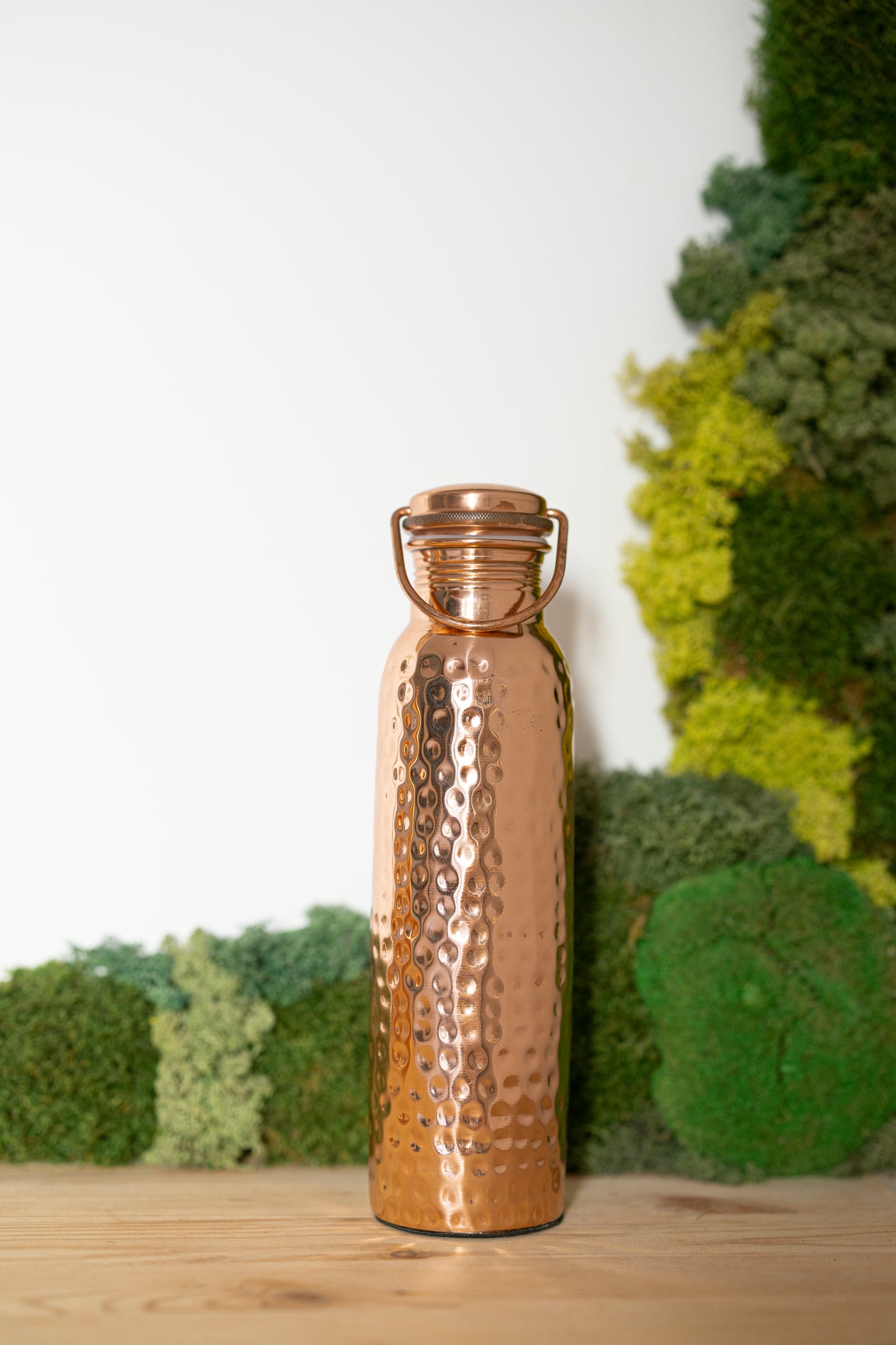 Botella de agua de cobre Kopero Martillada, 900 ml, de pie sobre una mesa de madera con un fondo de musgo