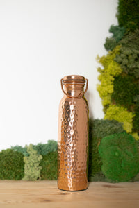 Botella de agua de cobre Kopero Martillada, 900 ml, de pie sobre una mesa de madera con un fondo de musgo