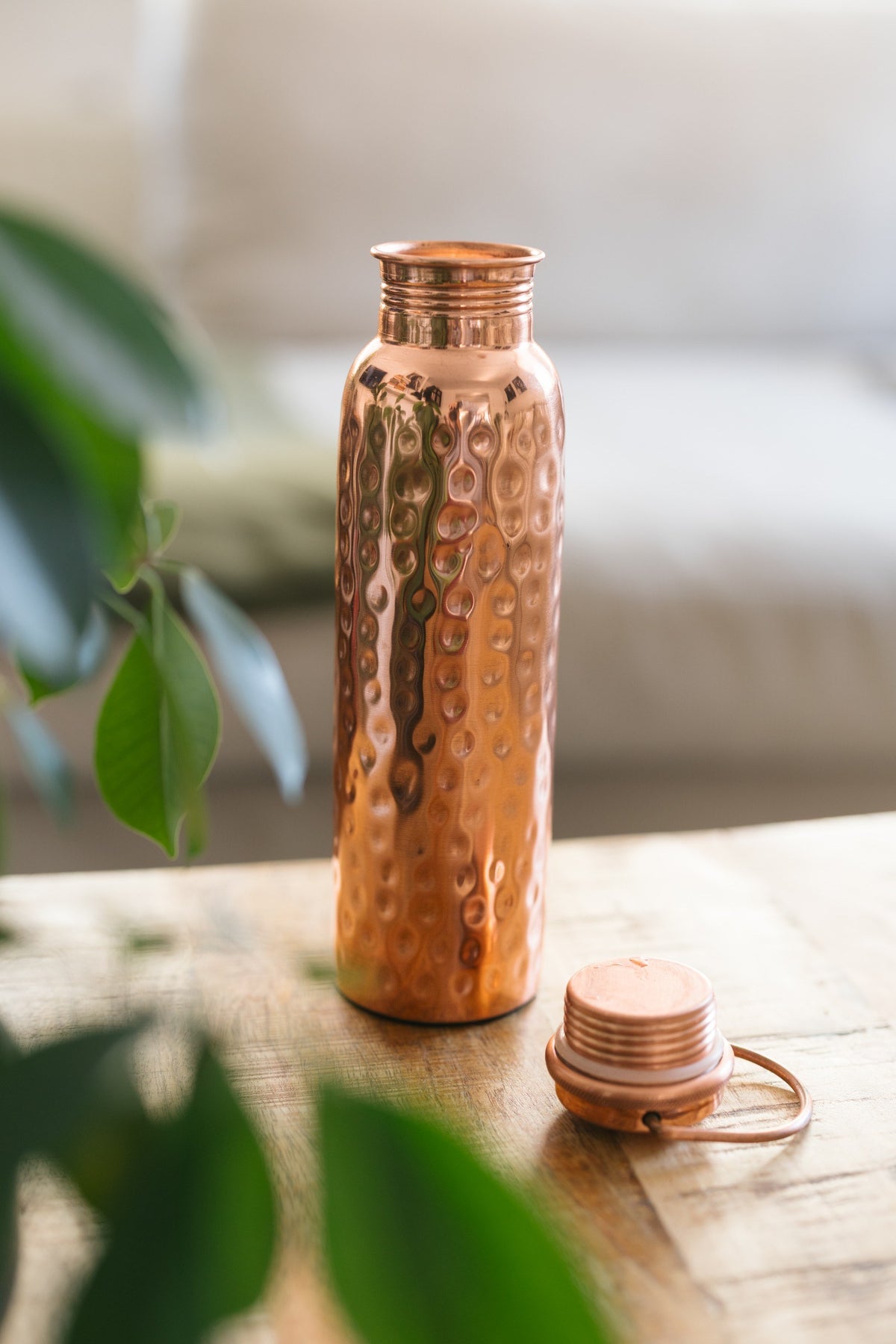 Botella de agua de cobre Kopero Martillado, de pie sobre una mesa de madera frente a un sofá, con el tapón suelto al lado