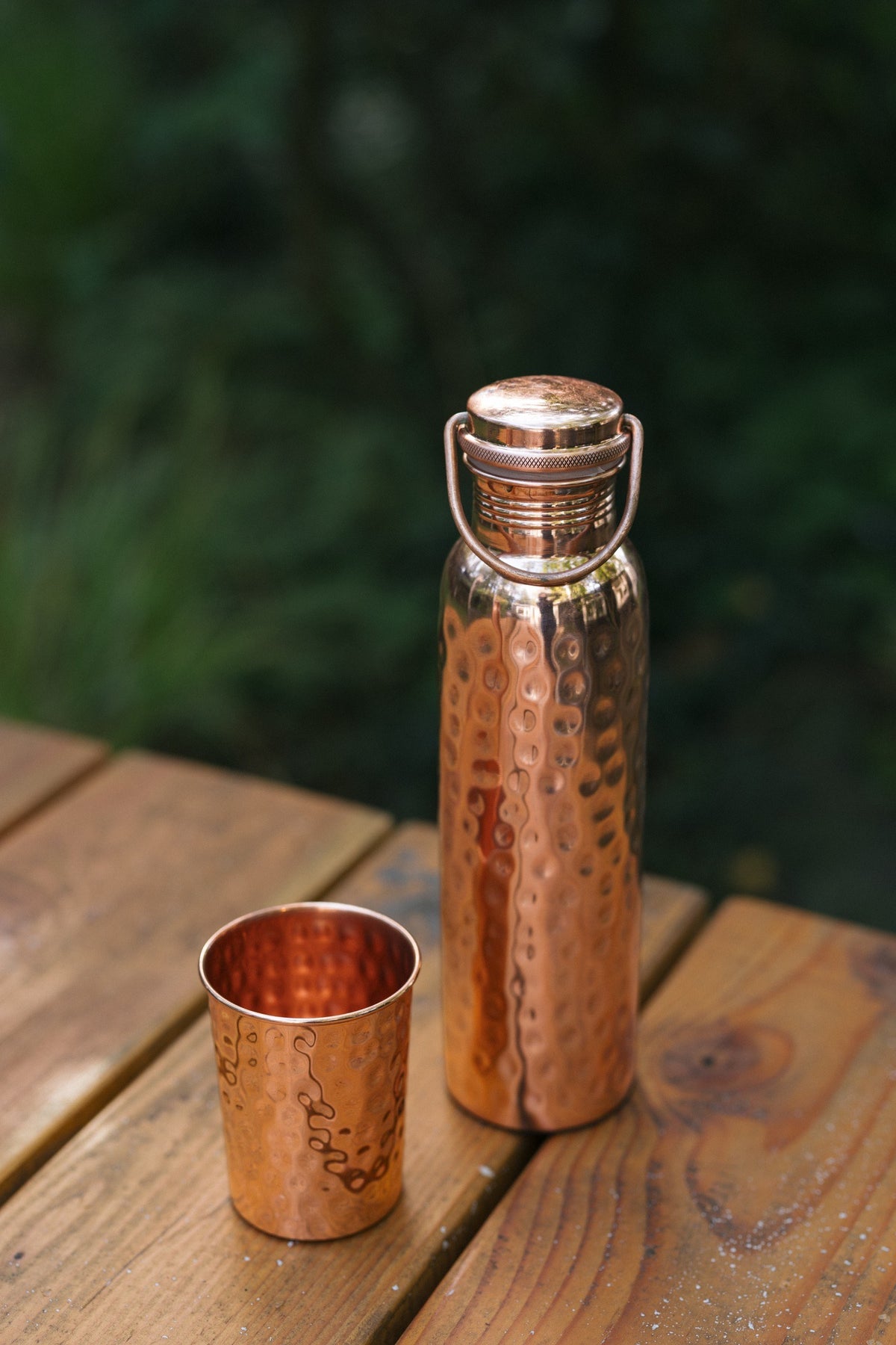 Botella de agua de cobre Kopero Martillado, sobre una mesa de jardín de madera junto a un vaso martillado