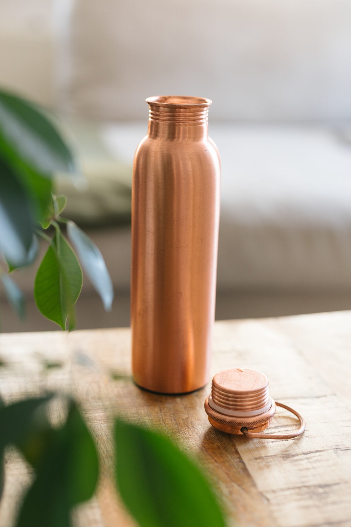 Botella de agua de cobre Kopero Liso, de pie sobre una mesa de madera frente a un sofá, con el tapón suelto al lado