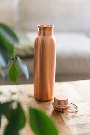 Botella de agua de cobre Kopero Liso, de pie sobre una mesa de madera frente a un sofá, con el tapón suelto al lado