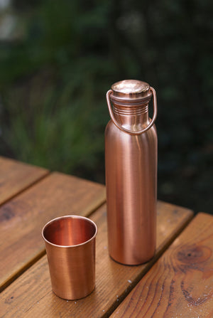 Botella de agua de cobre Kopero Liso, sobre una mesa de jardín de madera junto a un vaso de cobre