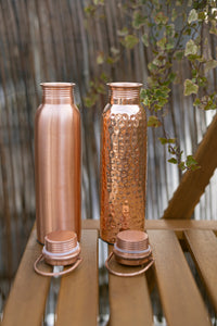 Dos botellas de agua de cobre Kopero, Lisa y Martillada, con el tapón abierto sobre una mesa de jardín de madera entre hiedra
