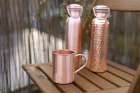 Botellas de agua de cobre Kopero Lisa y Martillada con una taza de cobre a juego sobre una mesa de jardín